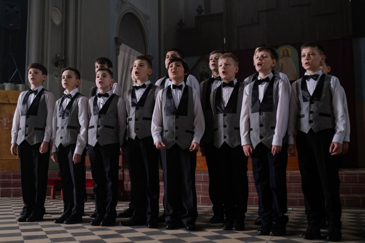 Group of boys singing in a choir, wearing formal uniforms in an indoor setting.