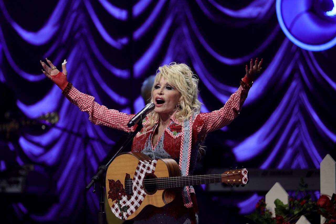 Dolly Parton performs with a guitar onstage, raising both arms as she sings into a microphone in a sparkling red-and-white outfit, with a dramatic purple-lit backdrop behind her. | Source: Getty Images