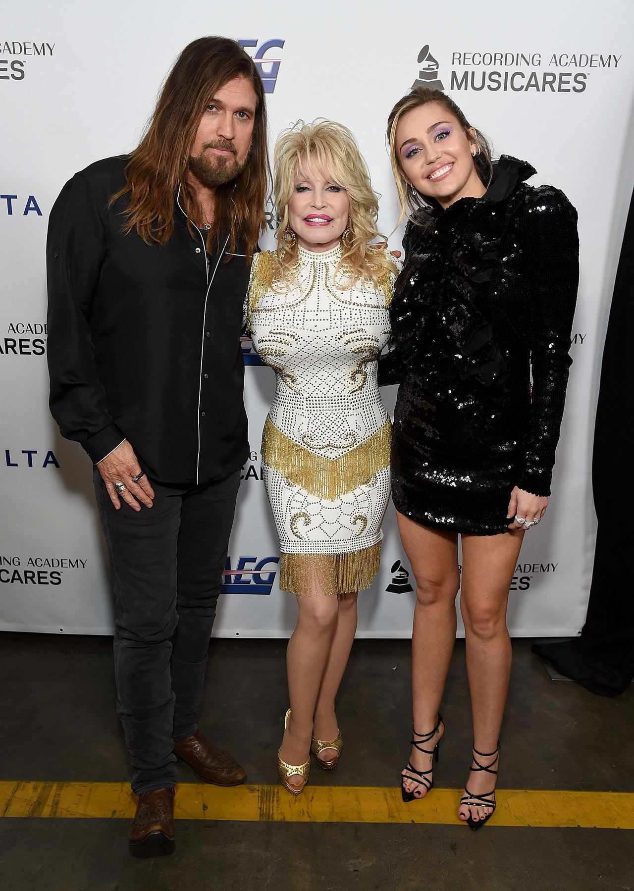 Billy Ray Cyrus, Dolly Parton, and Miley Cyrus pose together at the MusiCares Person of the Year event, with Parton standing between the father-daughter pair in a white-and-gold embellished outfit. | Source: Getty Images.