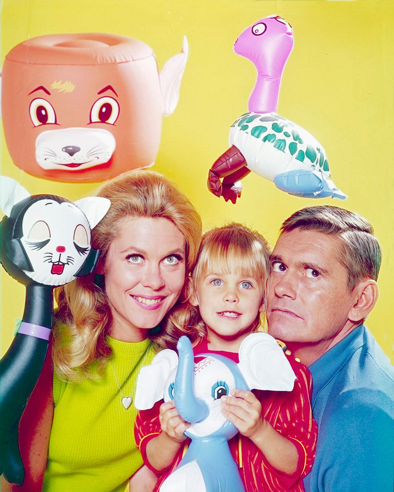 Erin Murphy poses between Elizabeth Montgomery and Dick York in a colorful "Bewitched" promotional image surrounded by oversized toy figures. Murphy is still a small child here, making the photo a sharp contrast with the polished selfies fans are seeing from her six decades later.