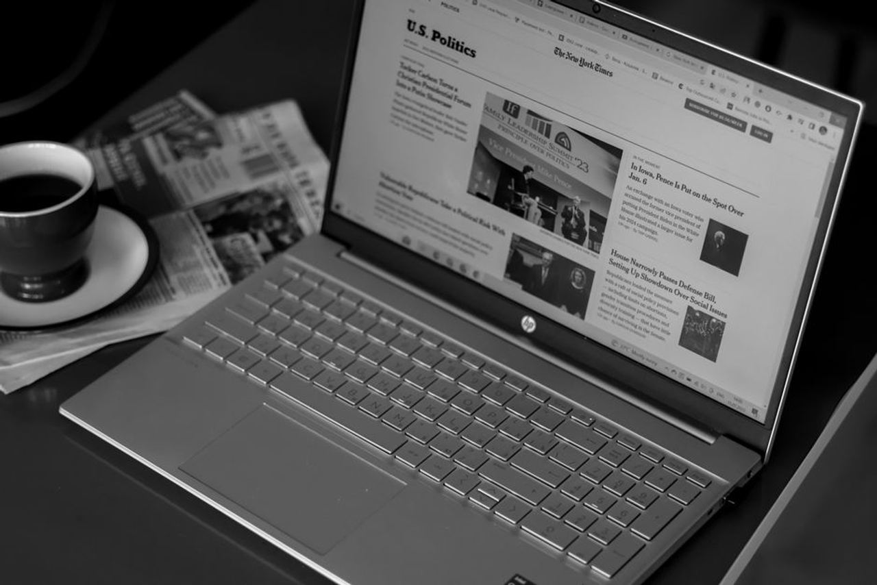Black and white image of a laptop displaying news articles, accompanied by a cup of coffee and newspapers.