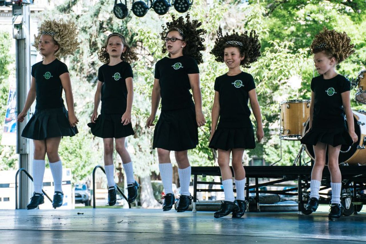 Young girls showcasing Irish dance on an outdoor stage, wearing wigs and traditional attire.