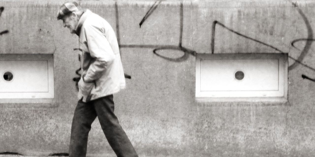 An elderly man walking alone along a worn wall, head down, lost in thought | Source: Flickr