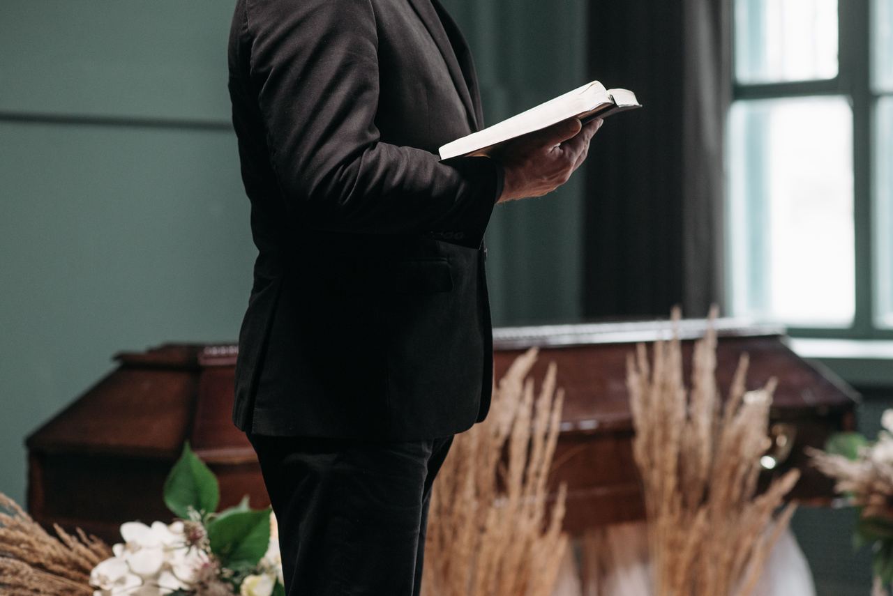 A pastor in a black suit holds a book during an indoor funeral ceremony. | Source: Pexels