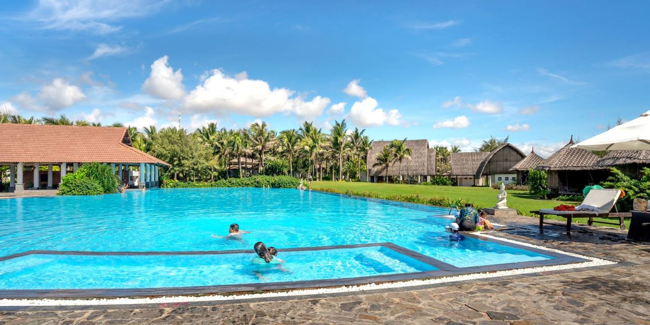 A large outdoor resort swimming pool surrounded by palm trees, landscaped gardens, lounge chairs, and traditional-style buildings | Source: Pexels