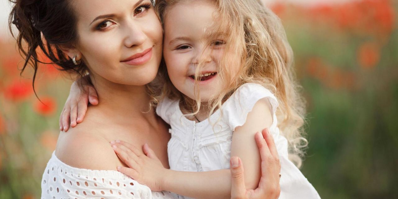 A smiling mother holding her young daughter close outdoors, both wearing white, sharing a warm embrace | Source: Magnific