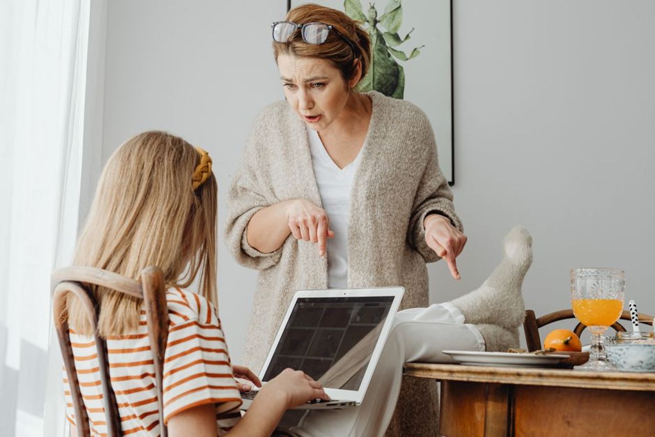 A mother scolds her teenage daughter using a laptop at home, highlighting a generational clash.