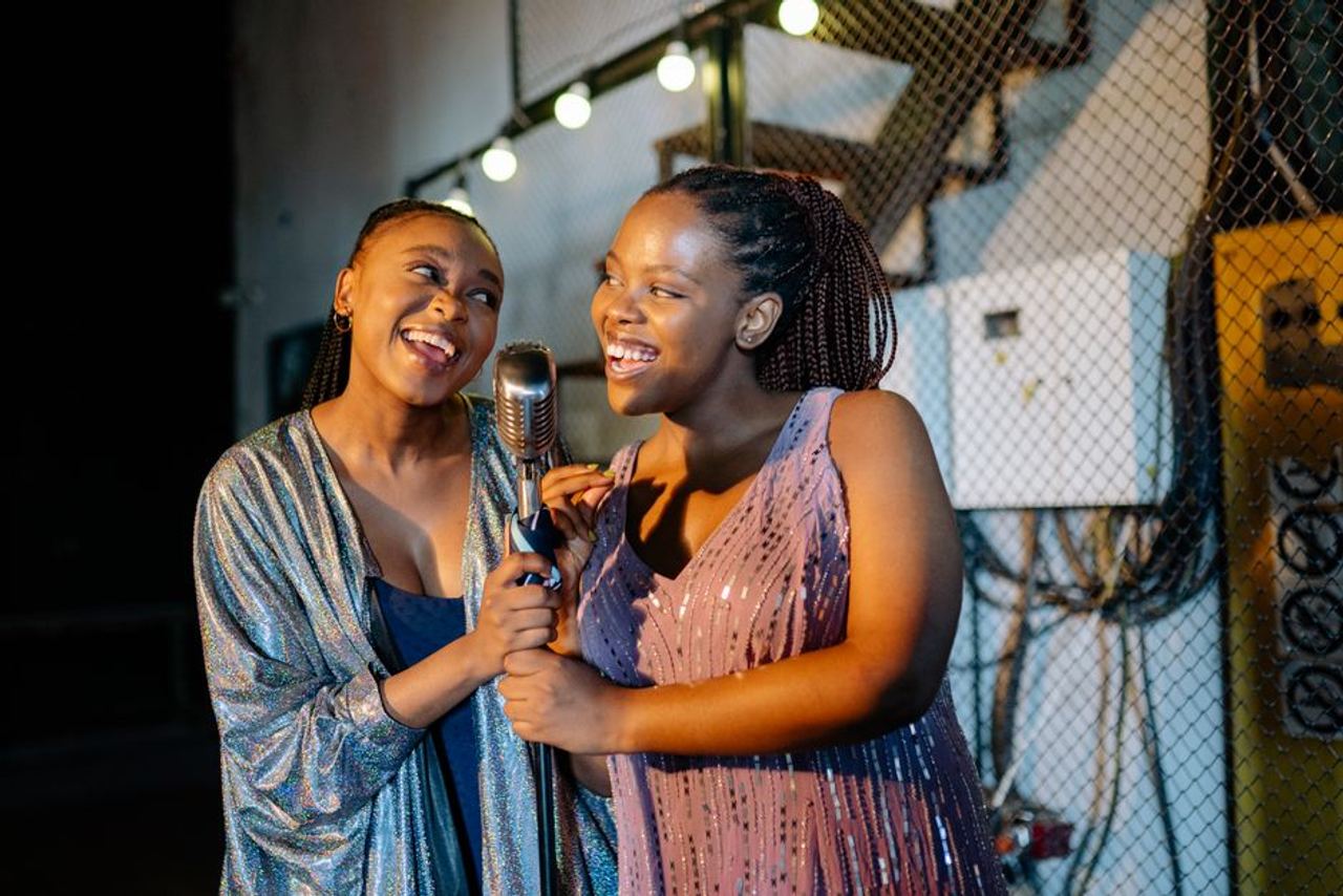 Two women singing joyfully on stage with a vintage microphone under warm lights.