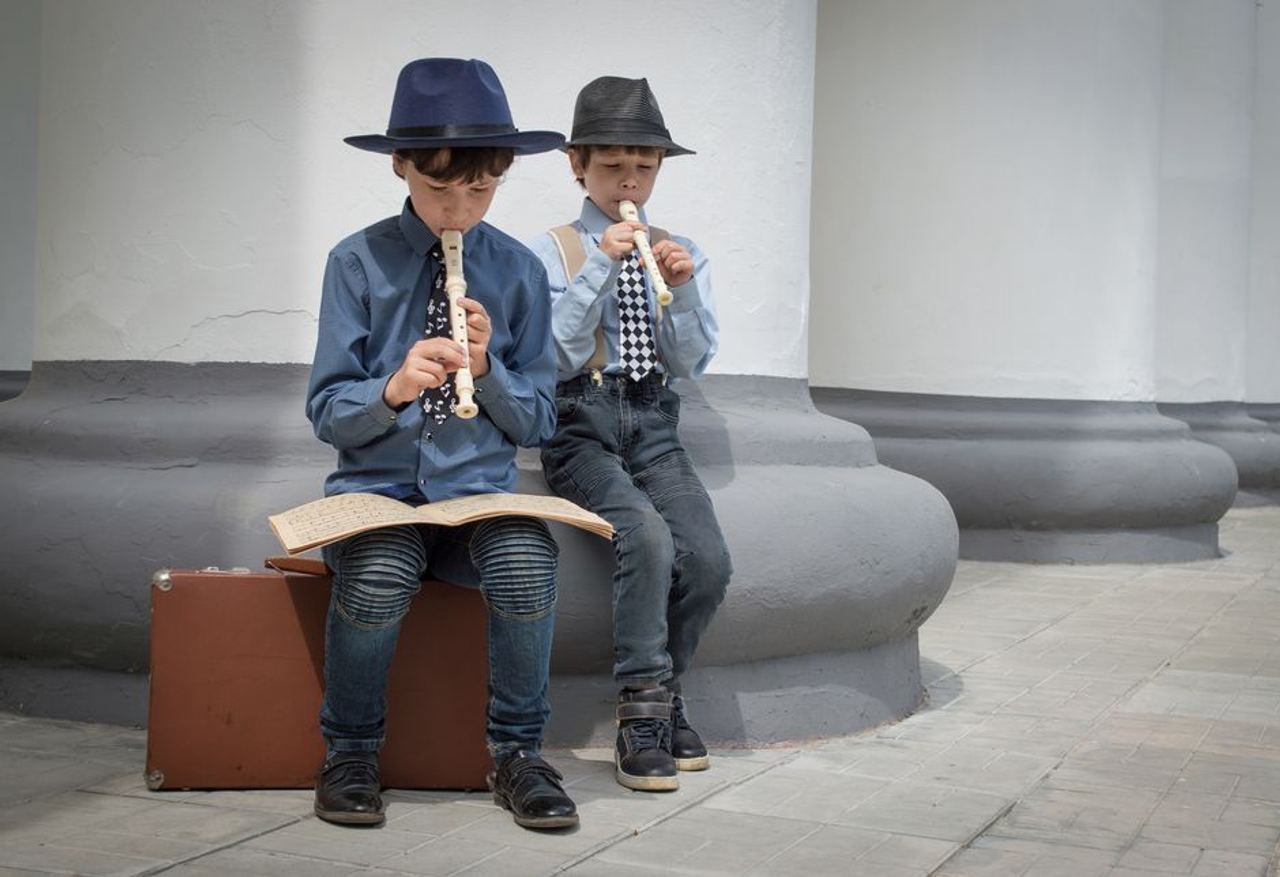 Two young boys in hats playing recorders outdoors with a music book, capturing youthful creativity.