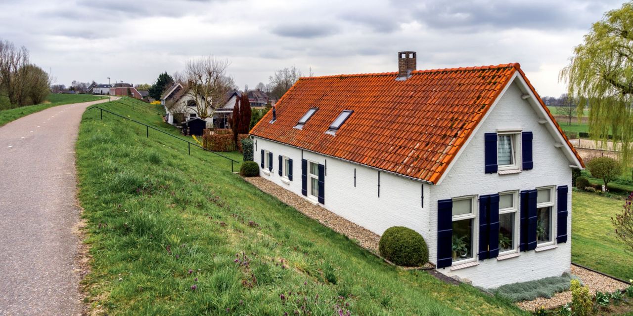 A white countryside house with a red-tiled roof, blue shutters, and a neatly landscaped yard beside a quiet rural road | Source: Magnific