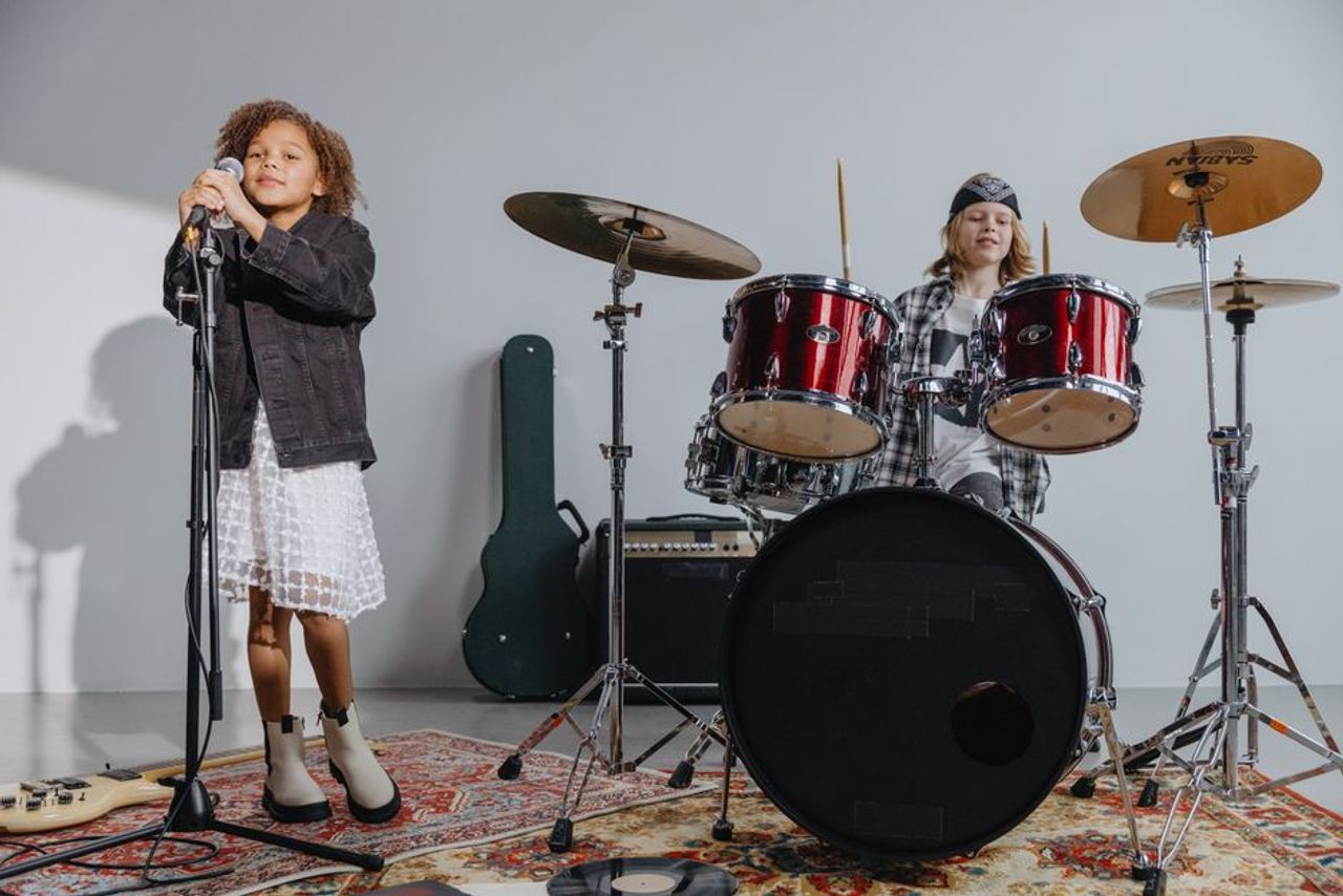 Two children performing in a music band with drums and microphone indoors.