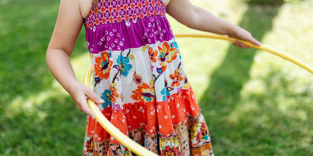 A young girl holding a yellow hula hoop while standing on a grassy lawn, wearing a colorful floral summer dress | Source: Pexels