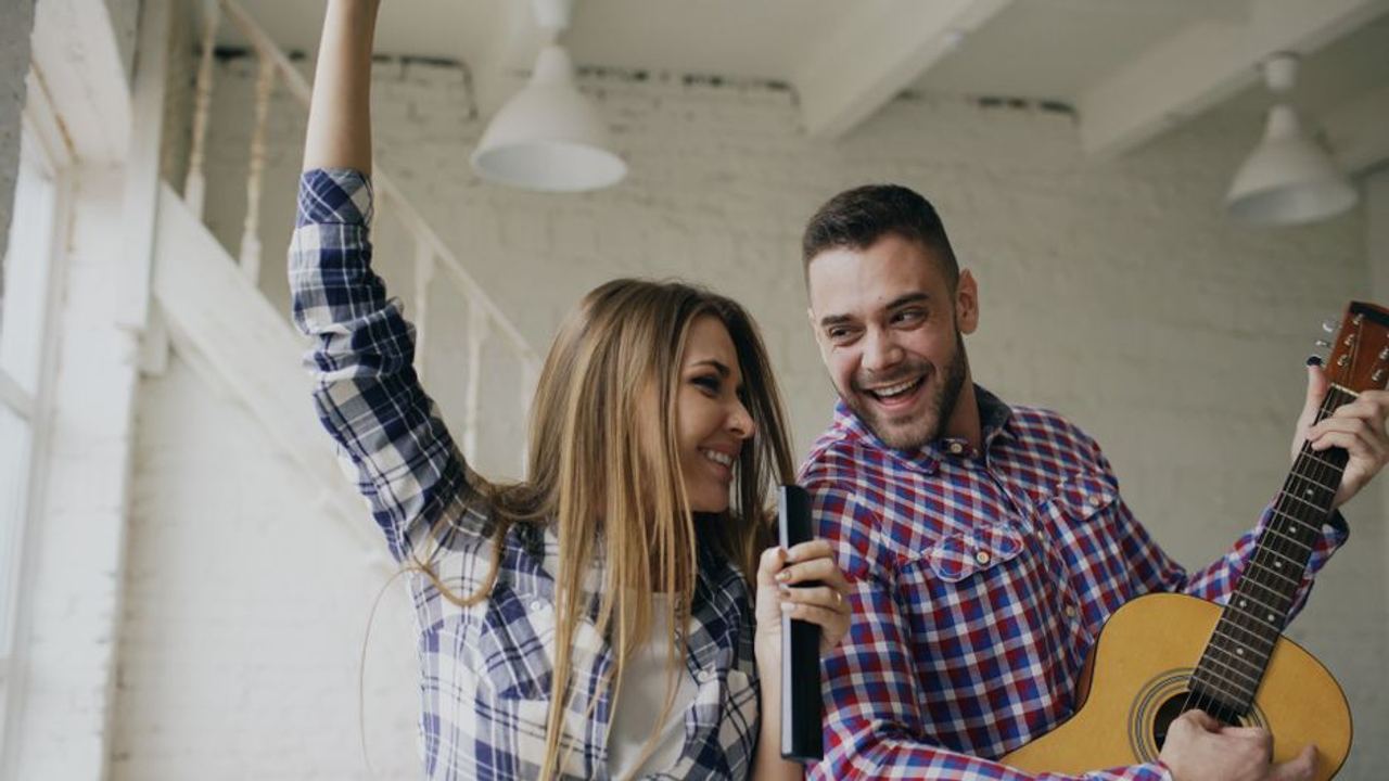 Cheerful couple having fun singing and playing guitar inside cozy room.