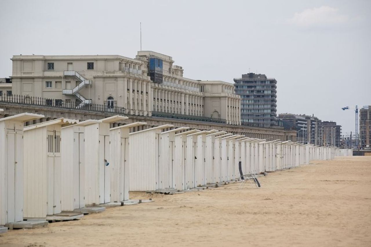 A row of beach sheds on a sandy Ostend beach with urban buildings in the background.