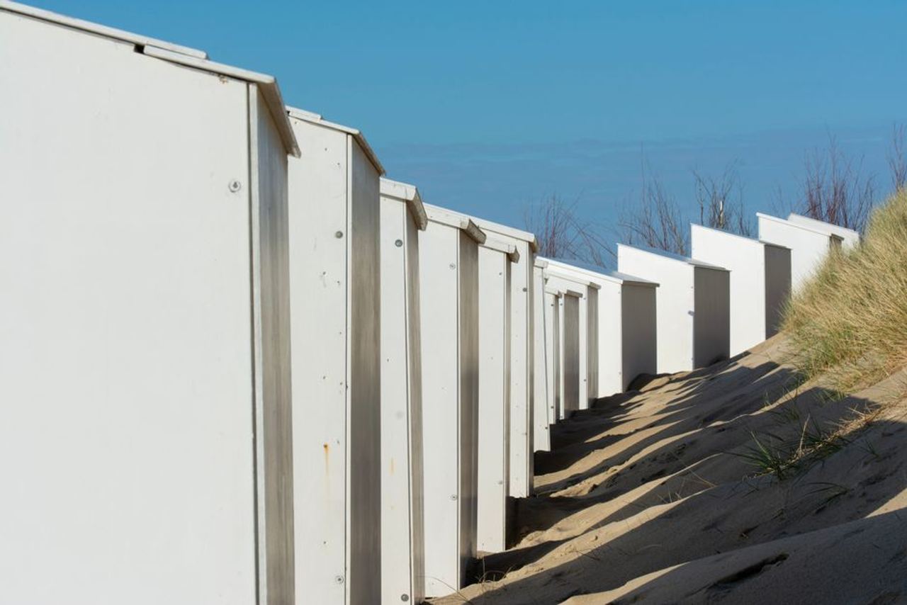 White beach huts line sandy dunes with shadows cast under the clear blue sky.