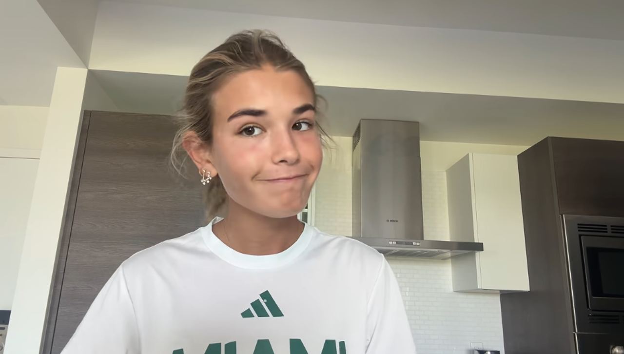 19-year-old Kai Trump flashed a playful grin in her modern kitchen during her September 5, 2026 video tour. Wearing a white Miami Adidas t-shirt and sparkling stud earrings, she stood before dark wood cabinetry, a stainless steel Bosch range hood, and a white tiled backsplash. | Source: YouTube/Kai Trump