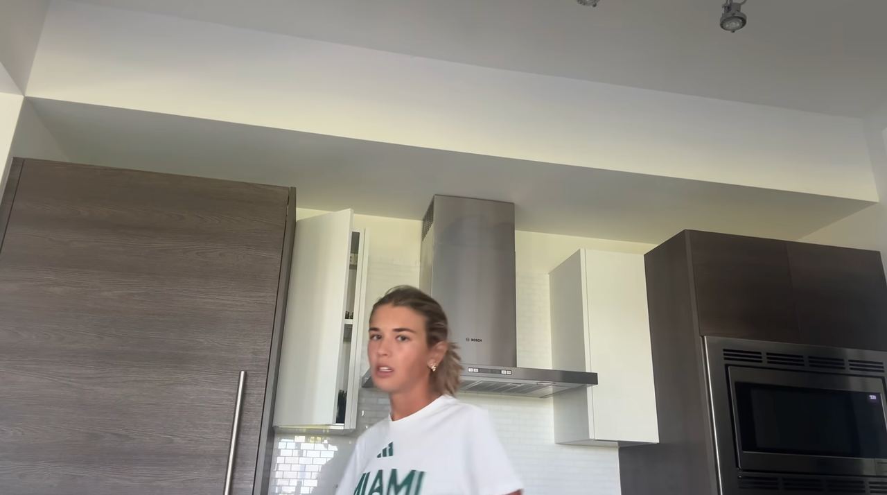 Kai Trump stands inside her kitchen wearing a white Miami t-shirt, framed by dark wood cabinetry, a stainless steel range hood, and built-in appliances under recessed ceiling lighting. | Source: YouTube/Kai Trump