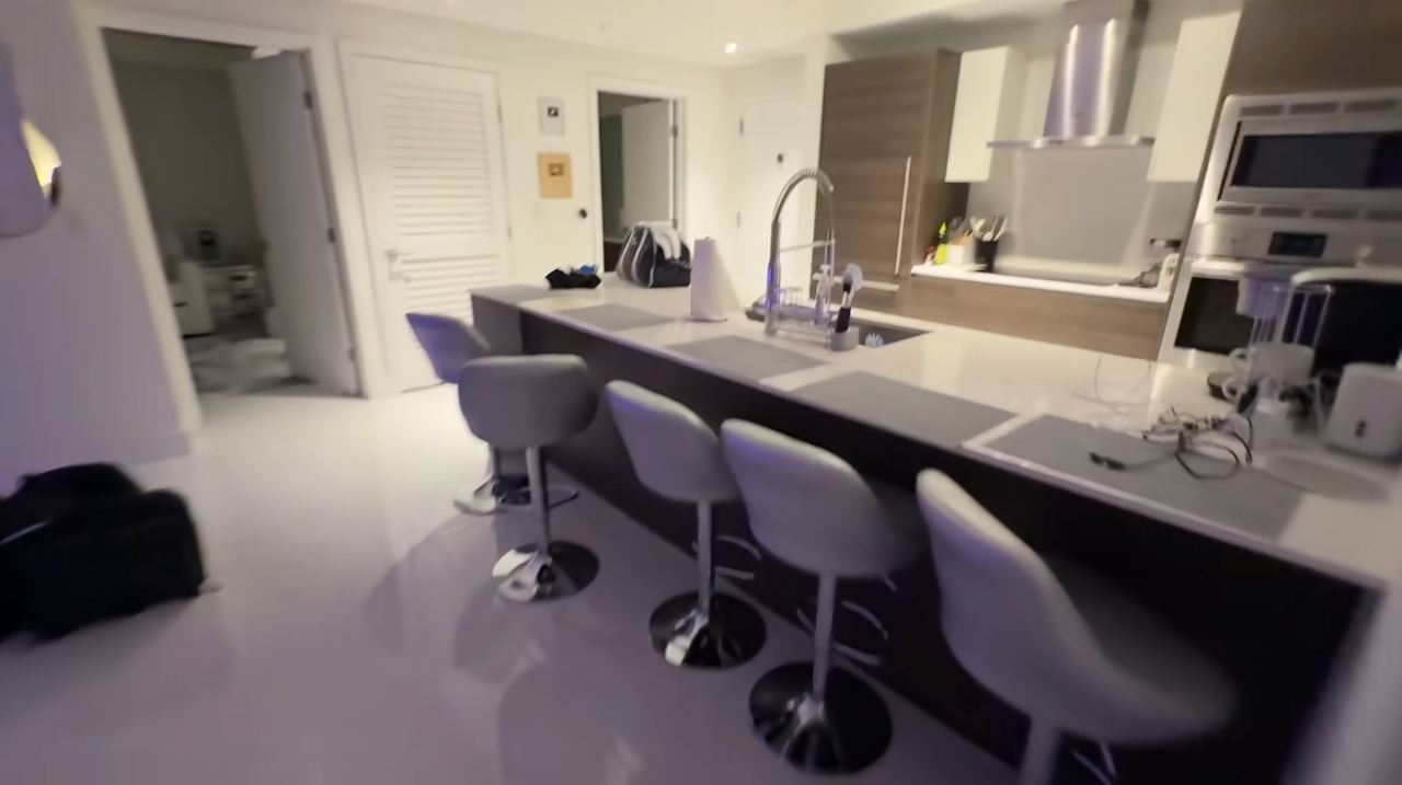 A modern kitchen peninsula features four gray leather barstools with sleek metallic pedestals, looking toward stainless steel built-in appliances and white quartz countertops. | Source: YouTube/Kai Trump