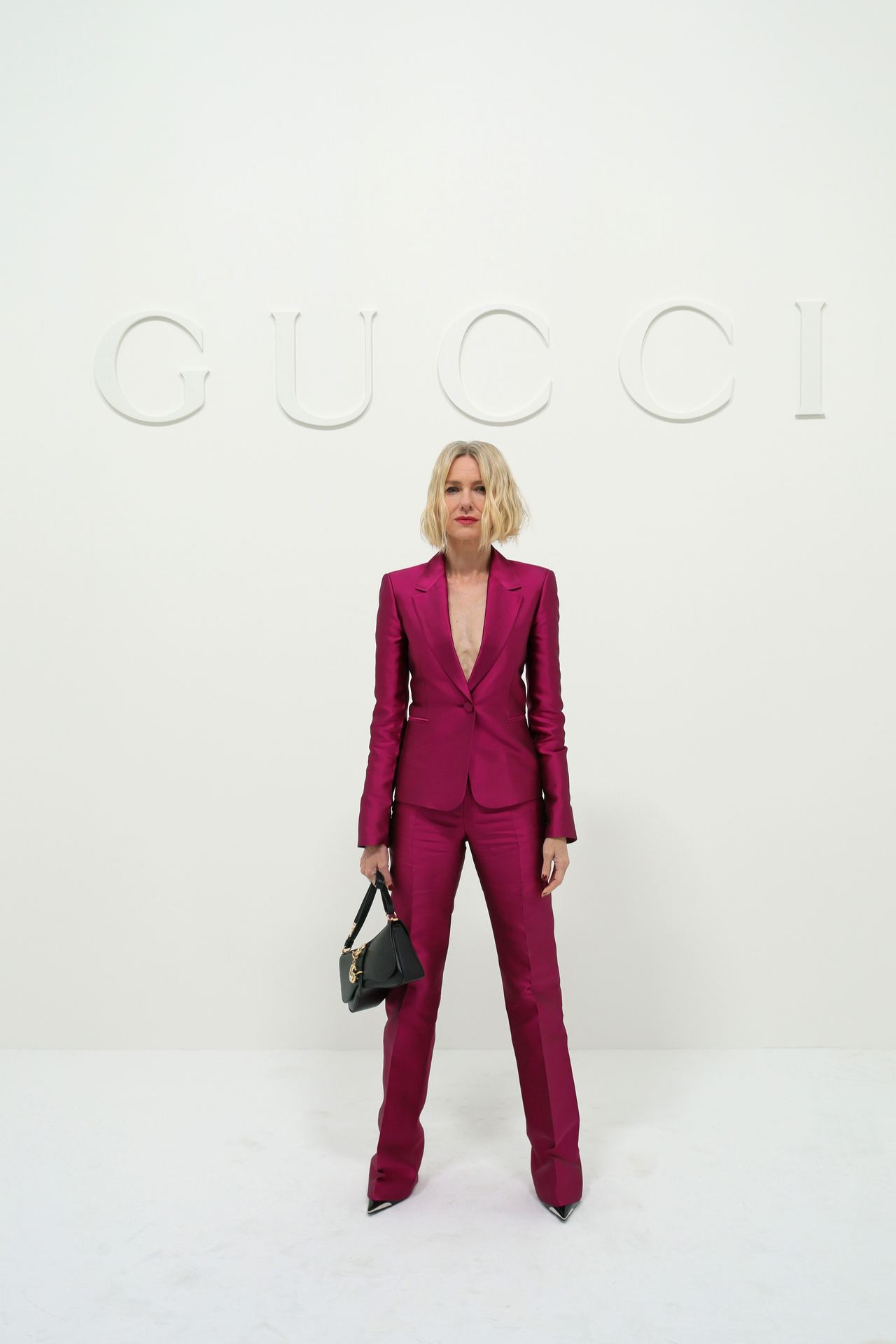 Naomi Watts commanded the minimalist photo wall beneath a subtle white Gucci logo at Allianz MiCo on September 25, 2026, during Milan Fashion Week Womenswear Spring/Summer 2027 in Milan, Italy. Striking a confident pose with one hand carrying a structured black purse, she highlighted the sharp tailoring and rich fuchsia tone of her satin suit alongside textured, chin-length blonde locks. | Source: Getty Images