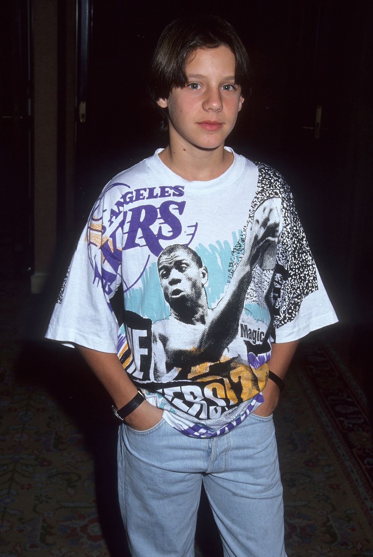 Omri Katz continues to grow into his teen years with a taller, leaner frame and chin-grazing dark curtain bangs that replace his early childhood neat cuts, wearing a graphic oversized Magic Johnson Los Angeles Lakers t-shirt paired with light-wash denim jeans. The photo was taken on July 27, 1991, as he attended the NBC Summer TCA Press Tour at the Universal Hilton Hotel in Universal City, California.
