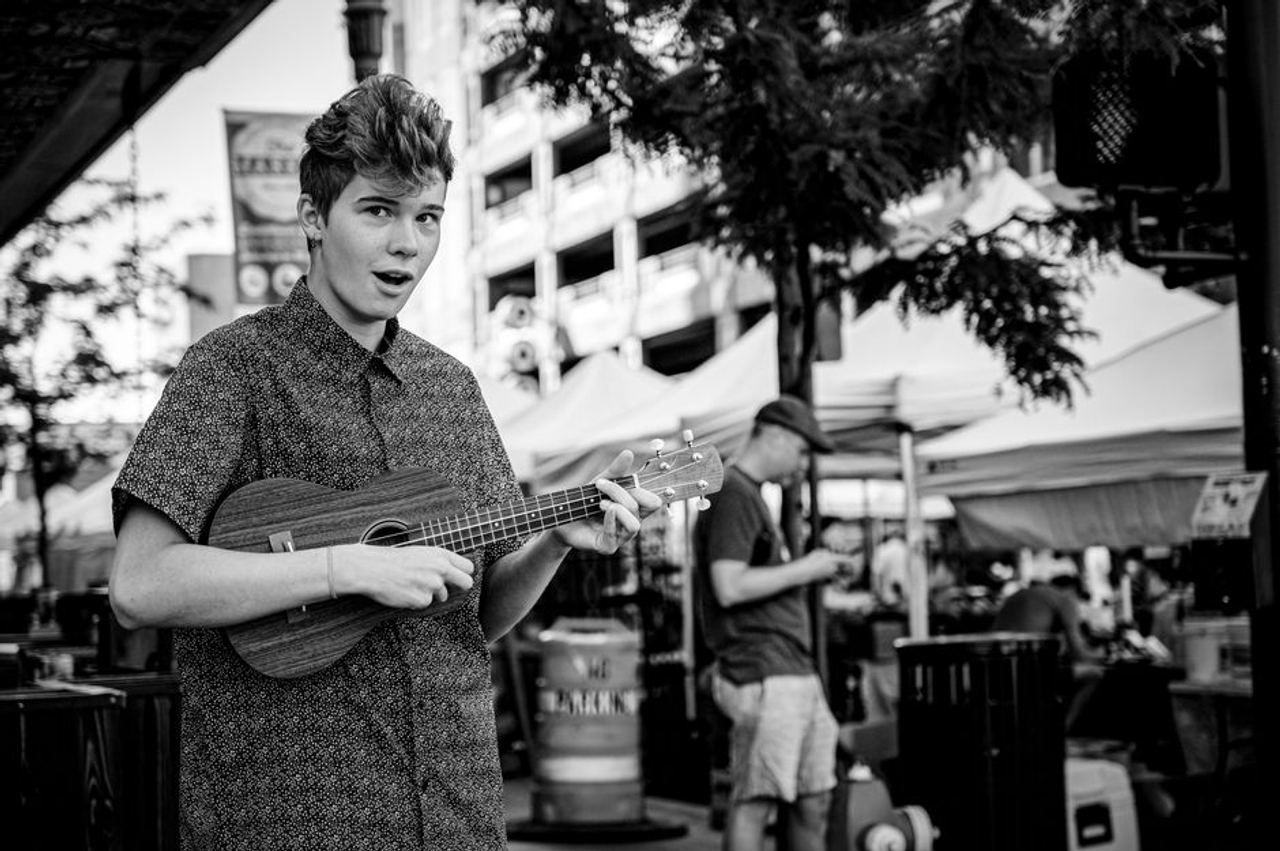 A young street musician playing a ukulele on a vibrant city street filled with outdoor market stalls.
