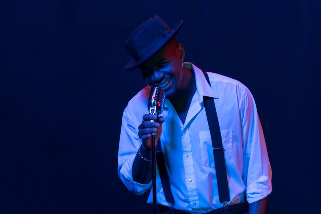 Black male jazz singer smiling while performing with vintage microphone and blue lighting.