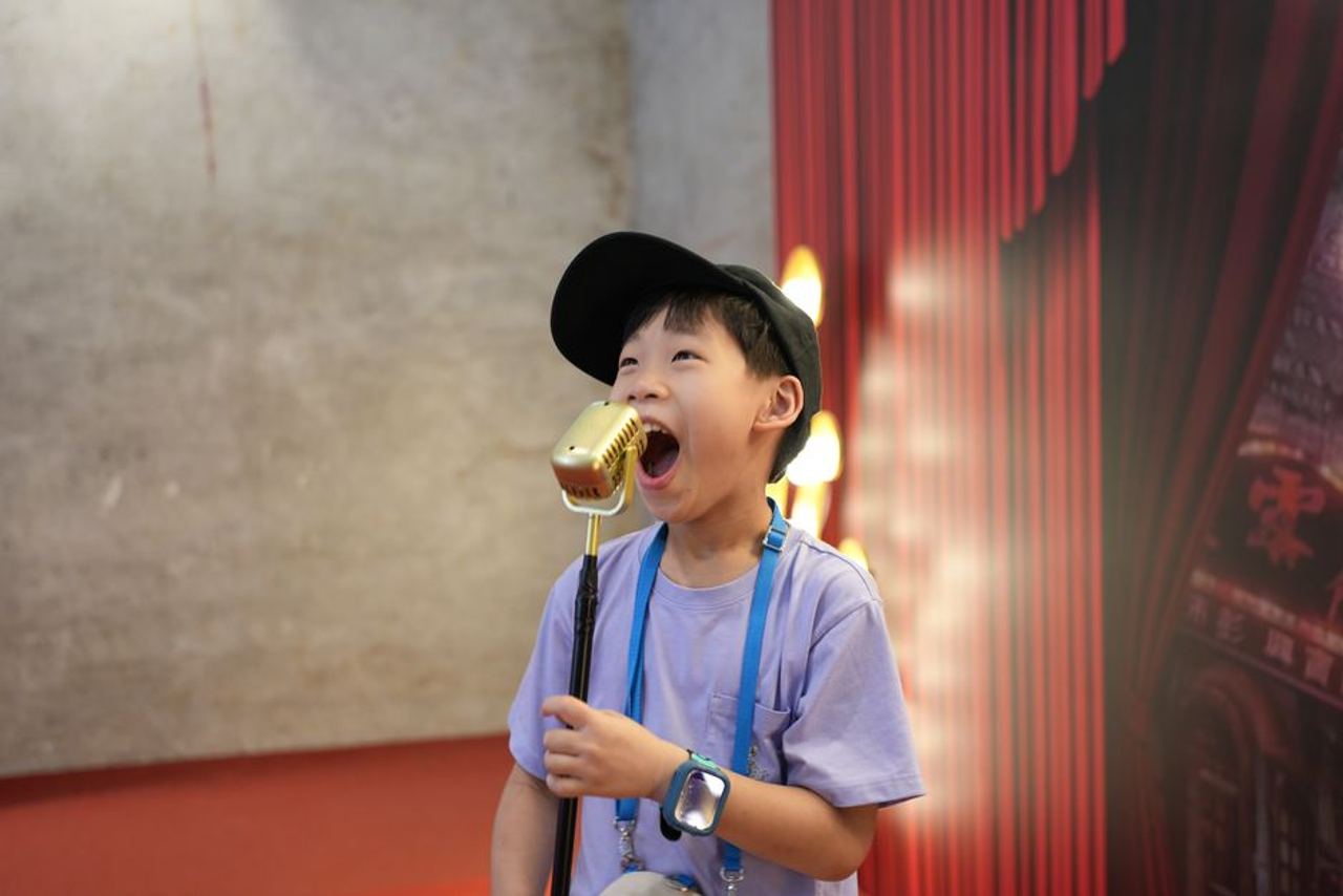 A young child passionately sings into a golden microphone on a stage with a red curtain backdrop.