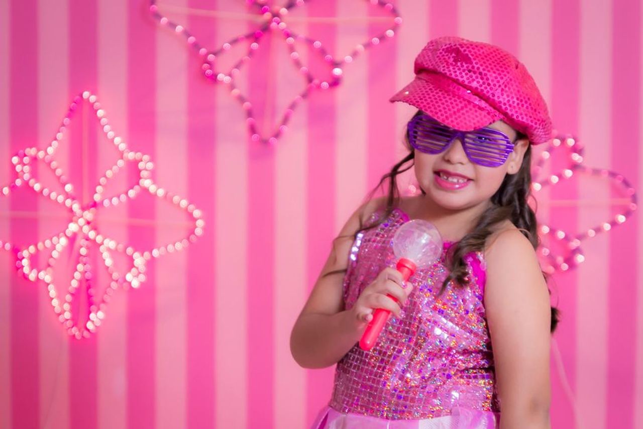 A cheerful girl in a pink sequined outfit and sunglasses poses with a toy microphone.