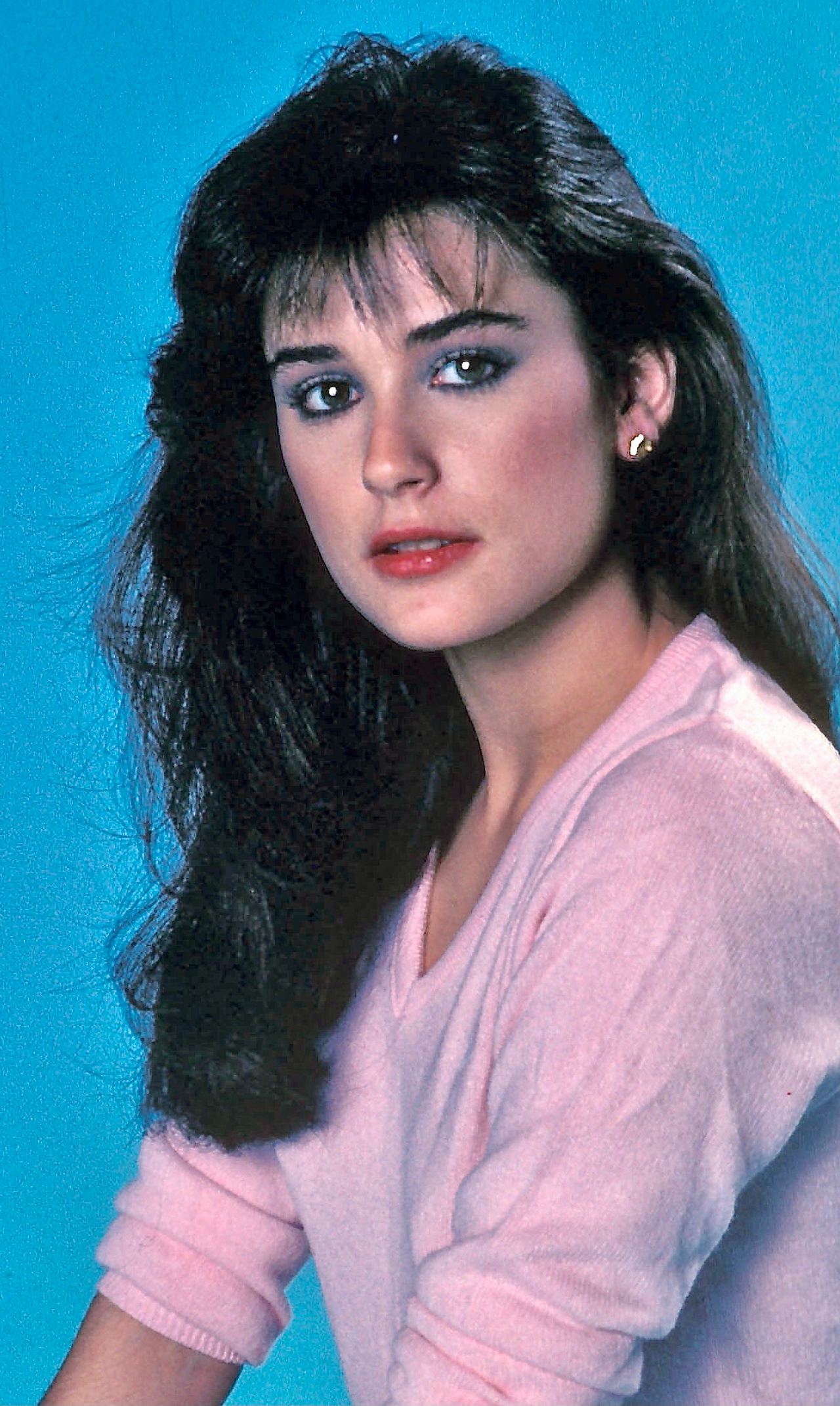 A young Demi Moore gazes softly into the distance, her dark, voluminous hair cascading over a pastel pink sweater that radiates warmth against a cool blue backdrop. Her expression is poised yet tender, framed by subtle makeup that highlights her natural charm.