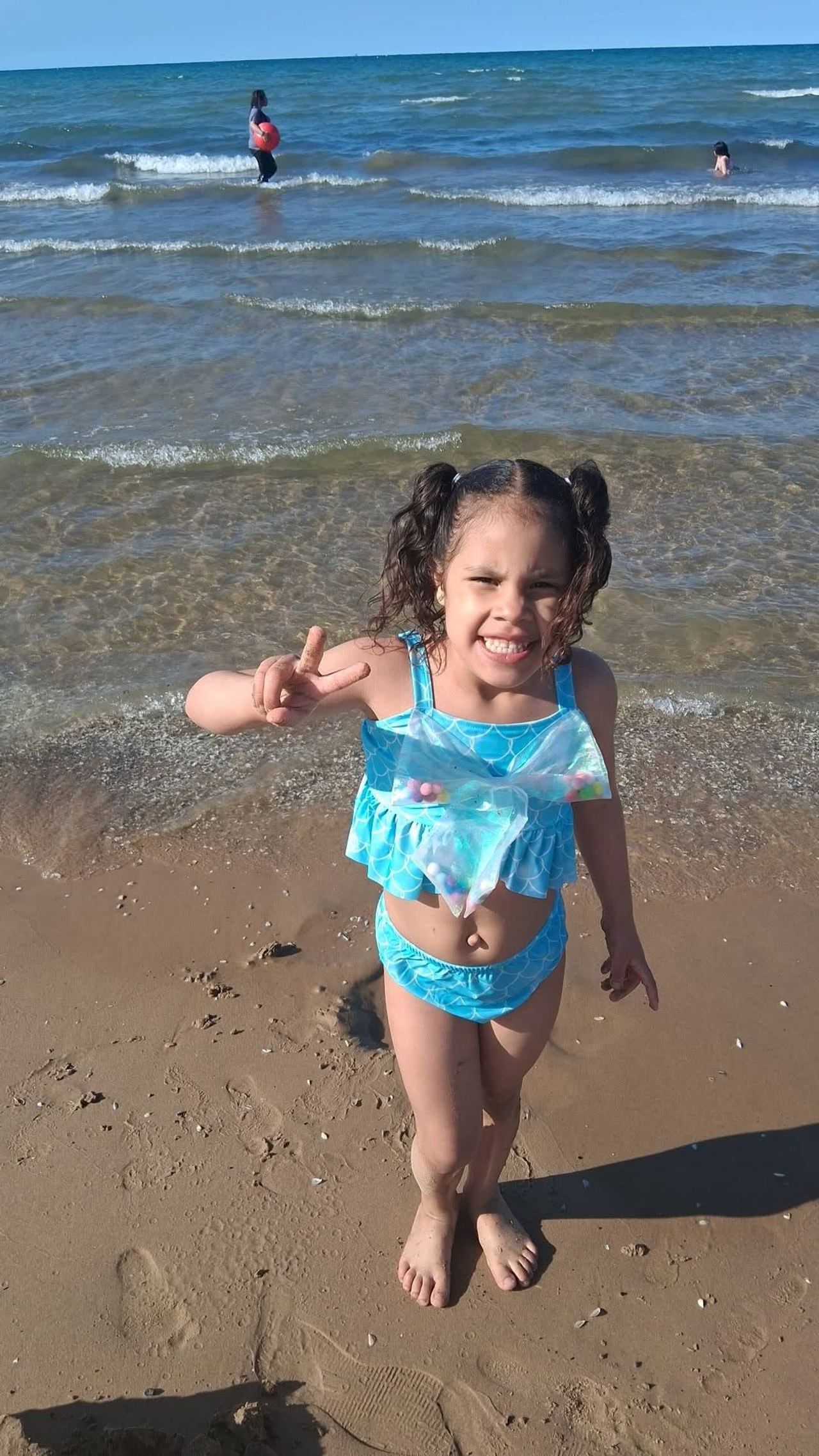 Eddimar Paola Hernández Chacón flashed a cheerful smile and held up a peace sign while standing barefoot on a sandy beach. Enjoying a sunny day by the water, she wore her hair in curly pigtails and sported a bright turquoise two-piece swimsuit accented with scale patterns, ruffles, and an iridescent bow. | Source: Facebook/Morena Esa