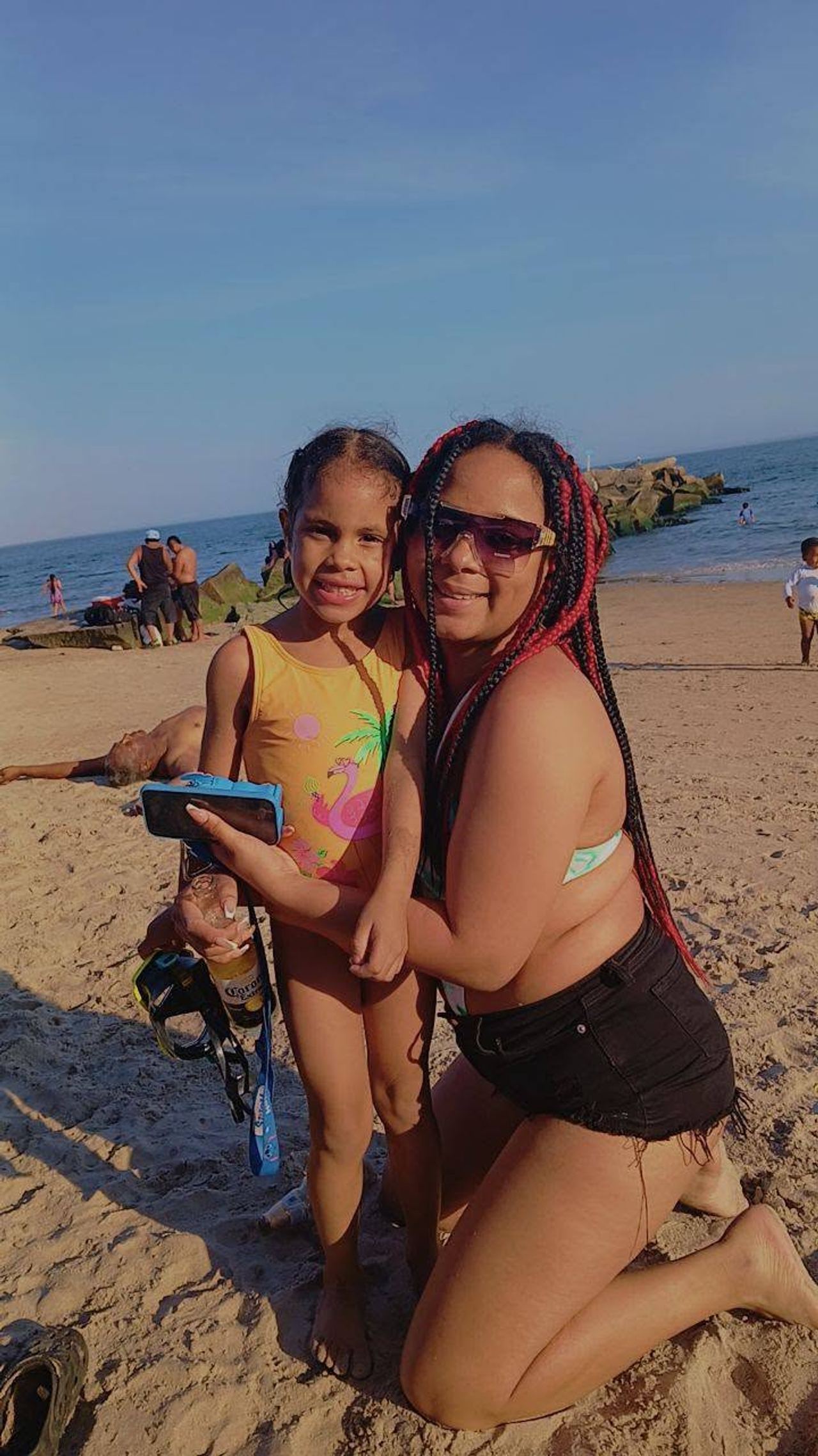 Eddimar Paola Hernández Chacón smiled happily while hugging her cousin on a sunny day at the beach. Eddimar wore a bright yellow swimsuit featuring a tropical flamingo graphic and styled her hair in braided pigtails, while her cousin knelt beside her on the sand wearing sunglasses, black denim shorts, and long red-and-black braids against the ocean waves. | Source: Facebook/Morena Esa