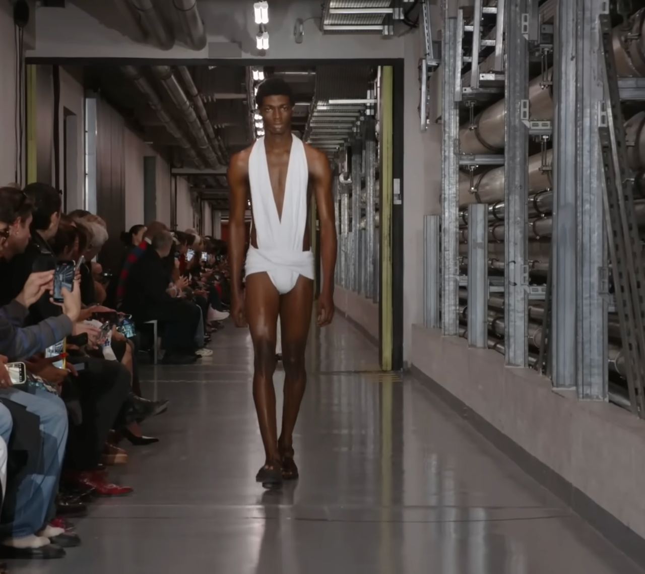 A male model in a cutout bodysuit design. This time in white, glowing strikingly against his dark chocolate skin. The look hinted that futuristic fashion might draw from the past, with a loin section reminiscent of the Roman subligaculum. From a post dated October 5, 2025. Source: Youtube/@jeanpaulgaultier