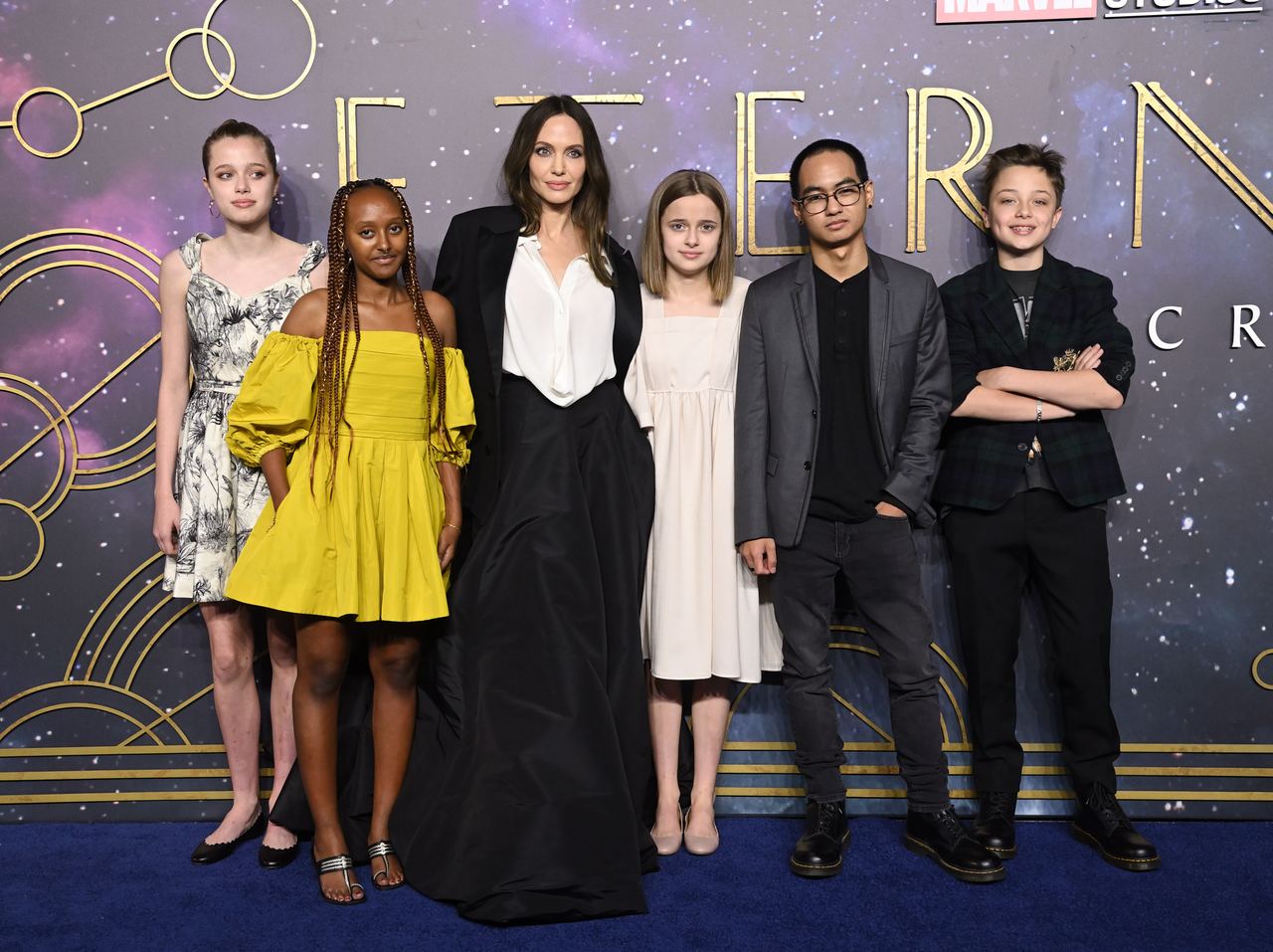 Angelina Jolie poses with her six children at the European premiere of Eternals, wearing a black floor-length gown over a white blouse, while her children stand beside her in a mix of formal and casual outfits. The family gathered on the blue carpet for the 2021 event, years before renewed attention surrounding the children’s use of the Jolie surname. | Source: Getty Images