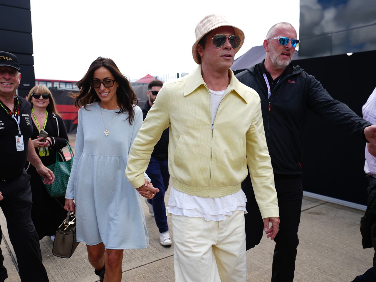 Brad Pitt and his partner Ines de Ramon walked hand-in-hand as they arrived at Silverstone Circuit in Northamptonshire on July 7, 2024. Pitt sported a light yellow zip-up jacket over a white ruffled hem, cream trousers, aviator sunglasses, and a houndstooth-patterned bucket hat, while de Ramon walked alongside him in a pastel blue long-sleeve shift dress, oversized dark sunglasses, and a pendant necklace. | Source: Getty Images