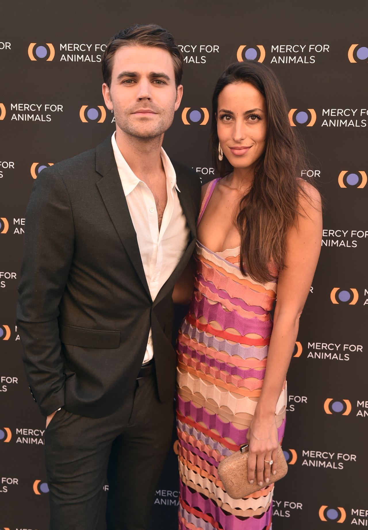 Paul Wesley and Ines de Ramon attended the Mercy For Animals 20th Anniversary Gala at The Shrine Auditorium on September 14, 2019, in Los Angeles, California. Wesley looked dapper in a tailored black suit paired with an open-collar white dress shirt, posing alongside de Ramon, who wore a colorful chevron-striped crochet slip dress in shades of purple, orange, and cream, accessorized with a sparkling clutch and delicate earrings against the event step-and-repeat. | Source: Getty Images