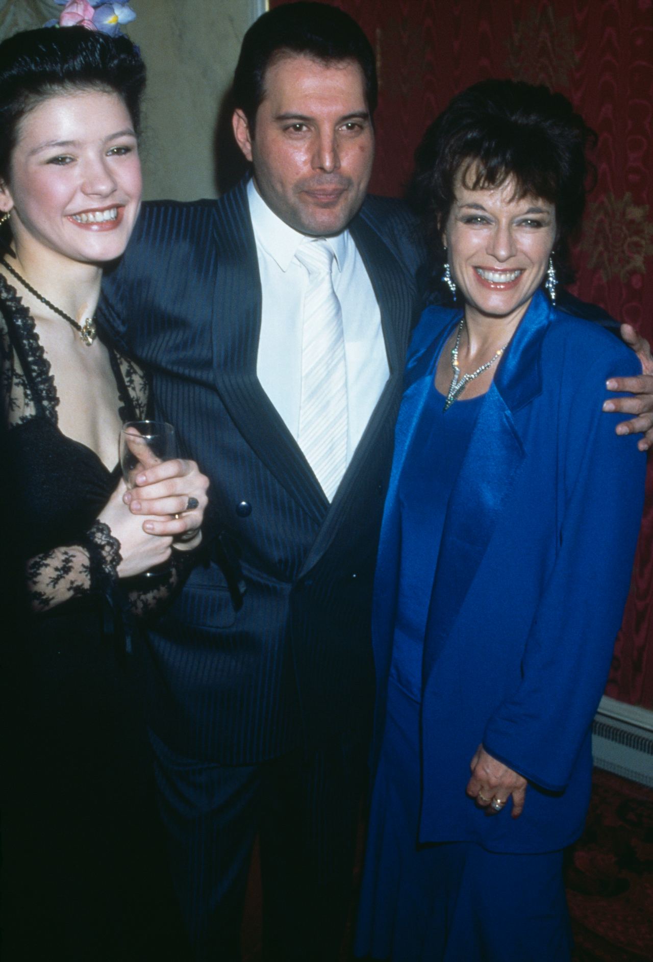 Catherine Zeta-Jones smiles radiantly while holding a glass of wine at a party for the new cast of the musical "42nd Street" on April 7, 1987, in London, England. The young actress wears an off-the-shoulder black dress with sheer lace long sleeves, a dark choker necklace, and floral hair accessories, posing alongside Queen frontman Freddie Mercury in a pinstripe double-breasted suit and actress Jill Gascoine in a bold cobalt blue jacket and dress ensemble.