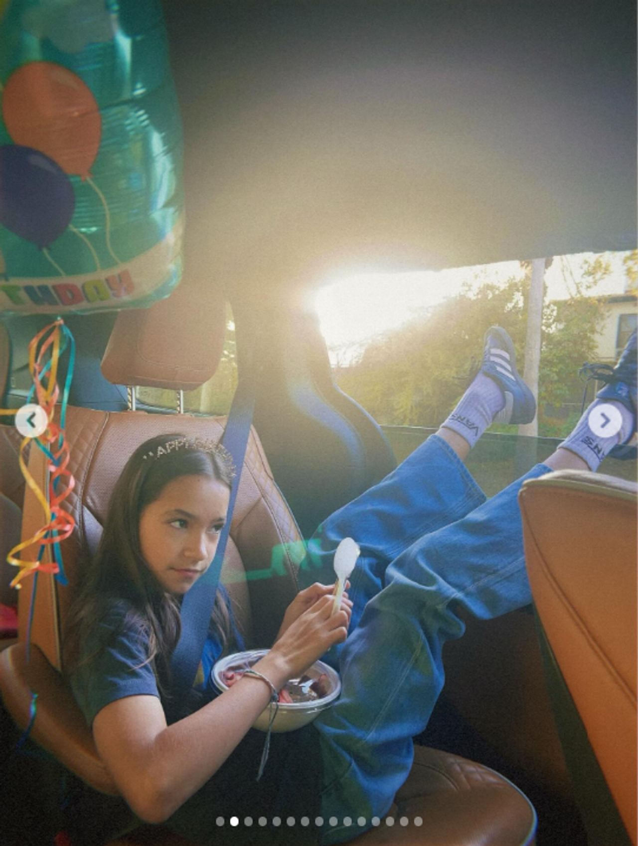 Fiona is pictured alone in the back seat of a car, wearing a dark blue T-shirt and blue jeans with white socks and sneakers. With her legs stretched across the seat, she holds a spoon over a small container of food in her lap, while sunlight streams through the window and colorful balloon decorations hang nearby. | Source: Instagram/mabellowe_