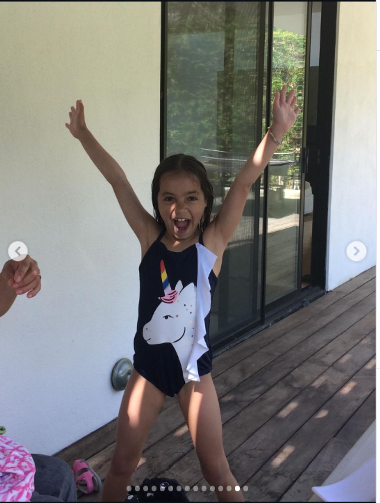 Fiona appears carefree in a playful childhood moment, standing on a wooden deck in a black one-piece swimsuit featuring a large white unicorn with a colorful horn. With both arms raised and a wide, exuberant expression, she looks full of energy as sunlight falls across the scene behind her. | Source: Instagram/mabellowe_