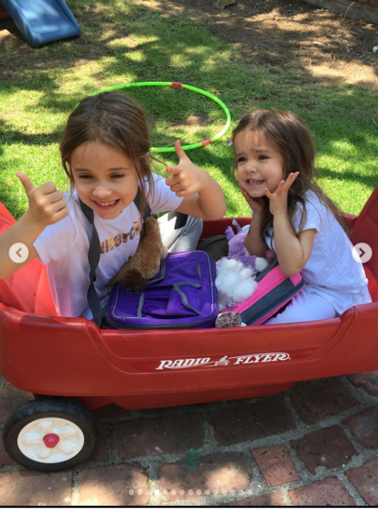 Mabel and Fiona share a playful moment in a large red Radio Flyer wagon, with Mabel giving two thumbs-up while Fiona smiles beside her. A purple bag and several toys sit between them, while a green hula hoop and children's slide are visible in the sunny backyard behind them. | Source: Instagram/mabellowe_