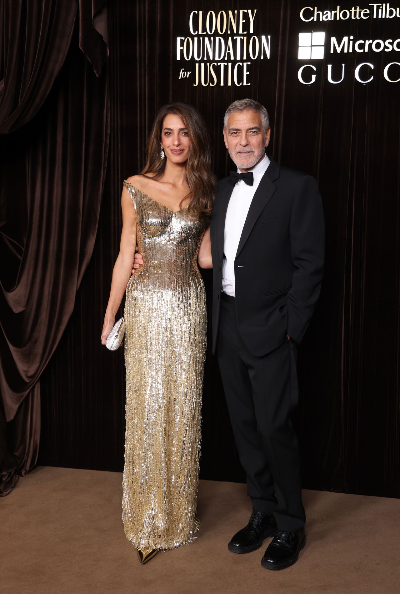 touches several generations of the family. George Clooney and his wife, Amal Clooney, pose together at The Albies, the annual awards ceremony hosted by the Clooney Foundation for Justice. Amal wears a shimmering gold, floor-length gown with an asymmetrical neckline and matching dangling earrings, while George looks sharp in a black tuxedo, white shirt, and bow tie. | Source: Getty Images