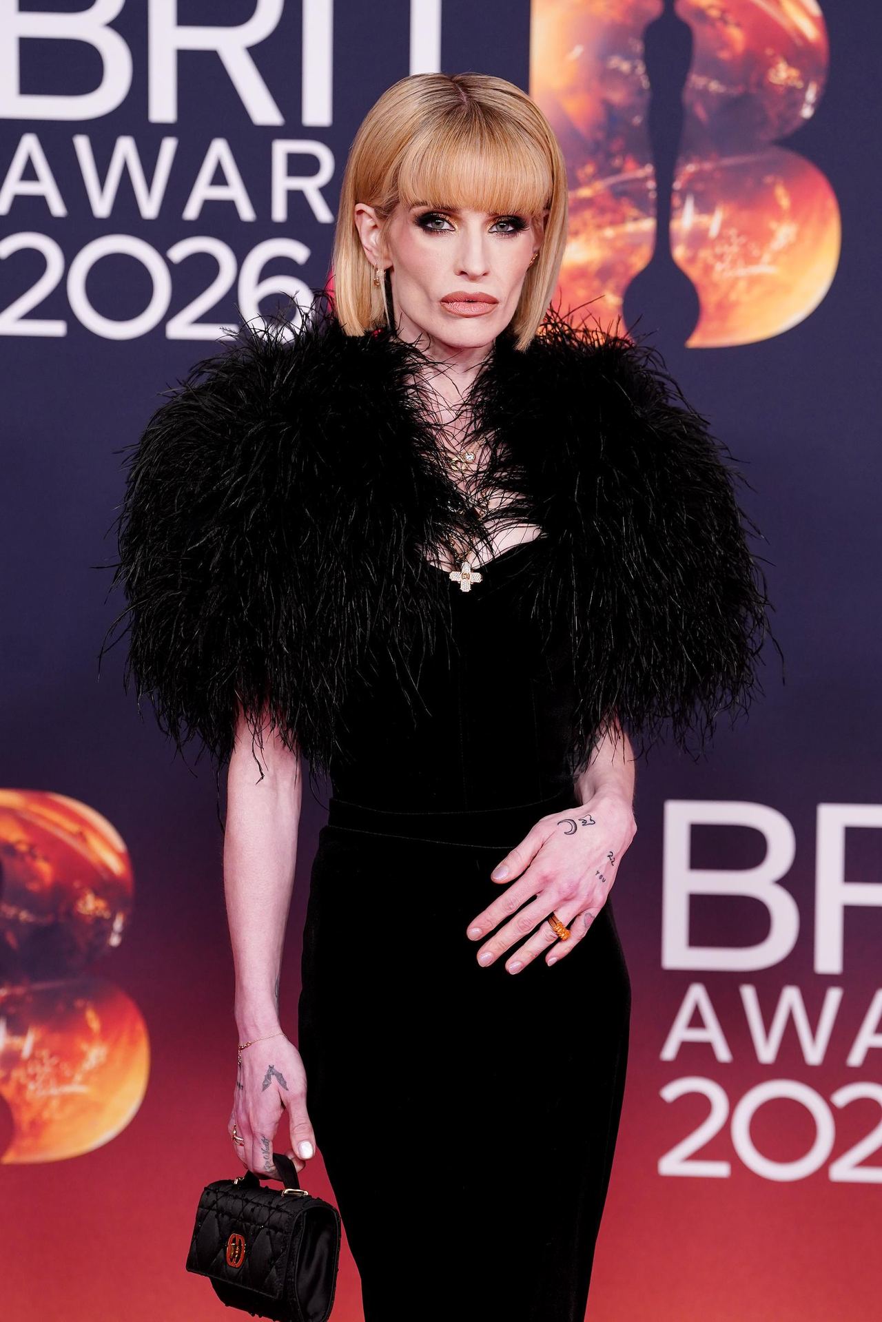 Kelly Osbourne wore a fitted black gown with dramatic feathered shoulders, a cross necklace, and a miniature handbag at the 2026 BRIT Awards in Manchester. | Source: Getty Images.