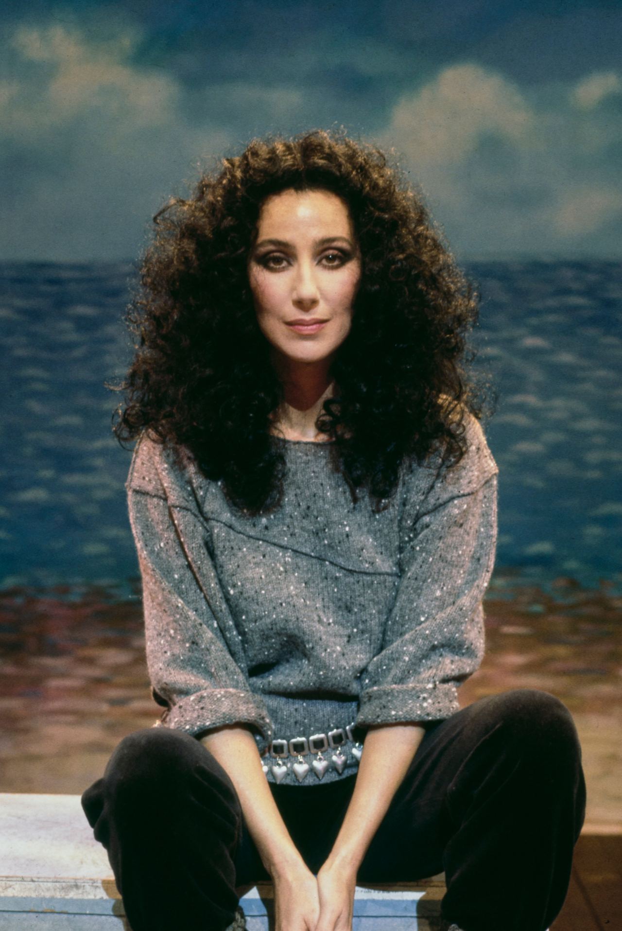Music and cultural icon Cher sat cross-legged with her hands resting between her knees in front of a painted beach backdrop in 1983. She framed her gaze with voluminous, cascading dark curls, dressed casually in a grey flecked knit sweater with rolled sleeves, accessorized with a metallic heart-charm belt over dark trousers. | Source: Getty Images