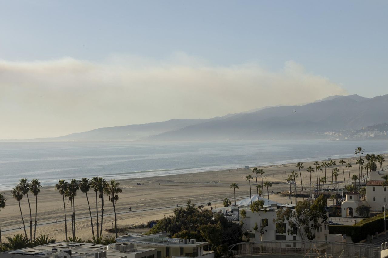 A wide elevated view captured Santa Monica Beach in Southern California on January 10, 2025, looking out over rows of tall palm trees, coastal structures, and the broad sandy shore toward the ocean. In the background, rolling coastal mountains stretched beneath a pale sky marked by a soft band of haze and smoke hanging over the horizon. | Source: Getty Images