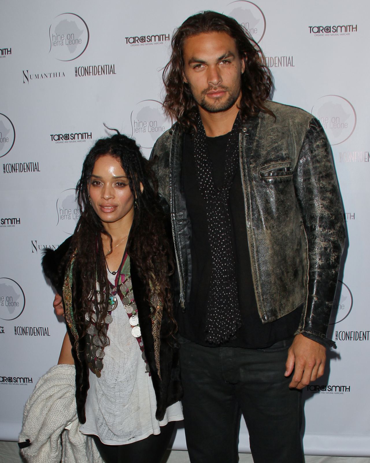 Jason Momoa and Lisa Bonet arrive at the Shine On Sierra Leone 5th Annual Fundraiser on May 25, 2011, in Venice, California. Momoa embraces Bonet while sporting shoulder-length wavy hair, a distressed leather motorcycle jacket, dark trousers, and a black dotted scarf draped over a dark tee, complementing Bonet's layered bohemian style featuring dreadlocks, an ornate coin necklace, and a dark vest over a flowing tunic. | Source: Getty Images