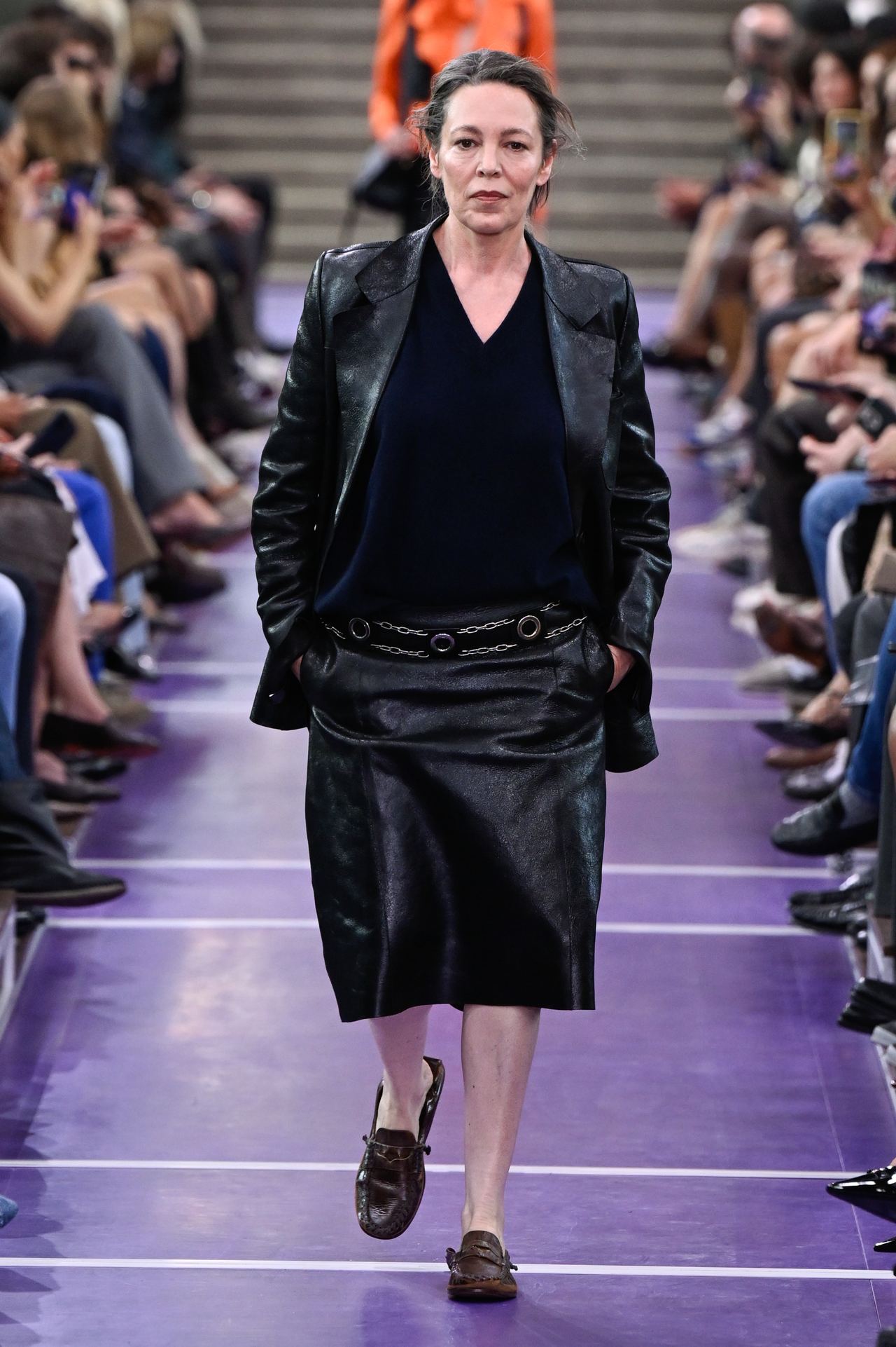 Olivia Colman walks the Miu Miu runway at the Palais d’Iéna during Paris Fashion Week, wearing a black leather blazer, navy top and knee-length leather skirt with brown loafers. | Source: Getty Images
