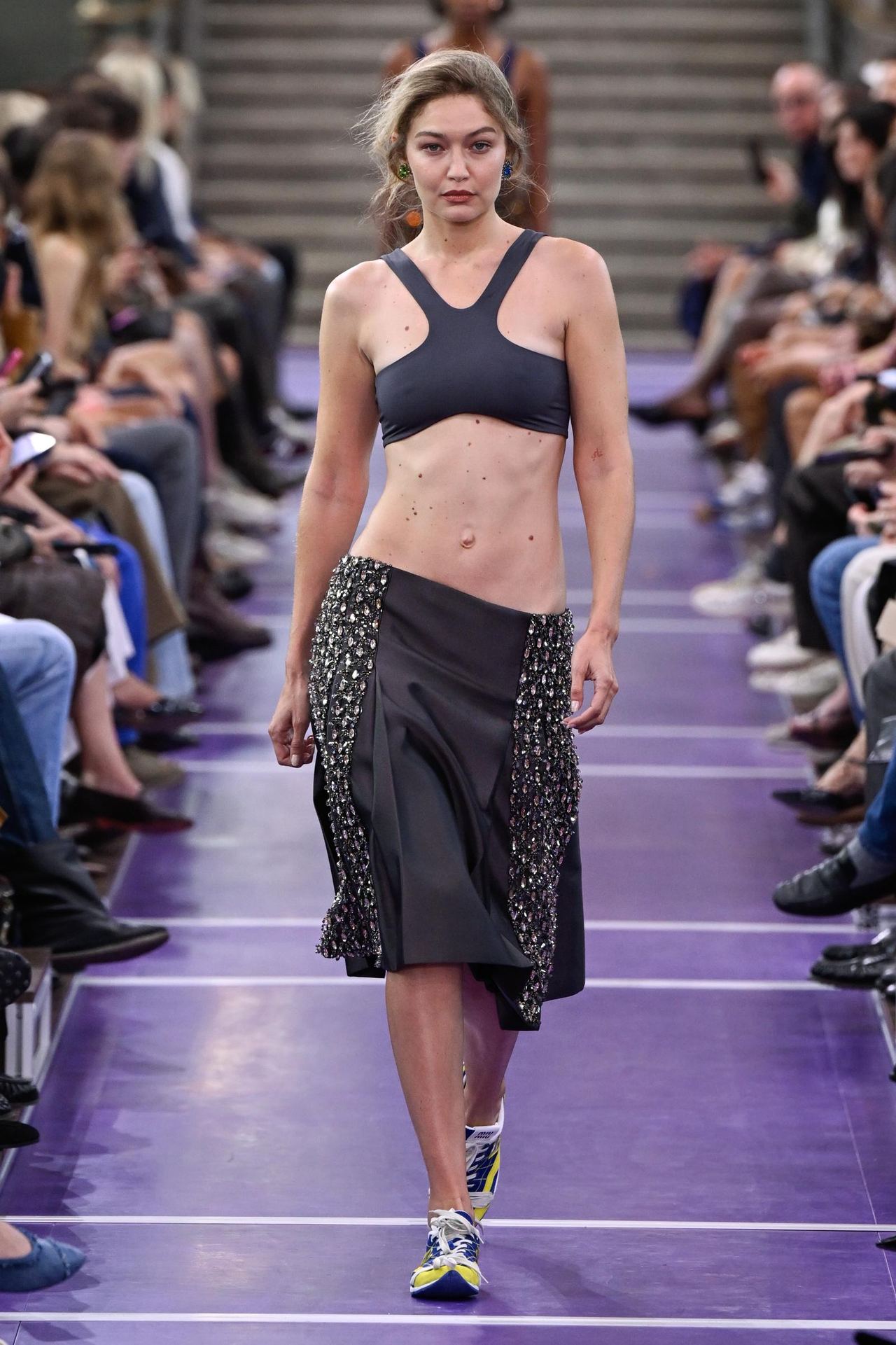 Gigi Hadid commands the Miu Miu runway in a charcoal cropped top and matching draped midi skirt, completing the look with colorful sneakers and her blonde hair swept loosely back. | Source: Getty Images