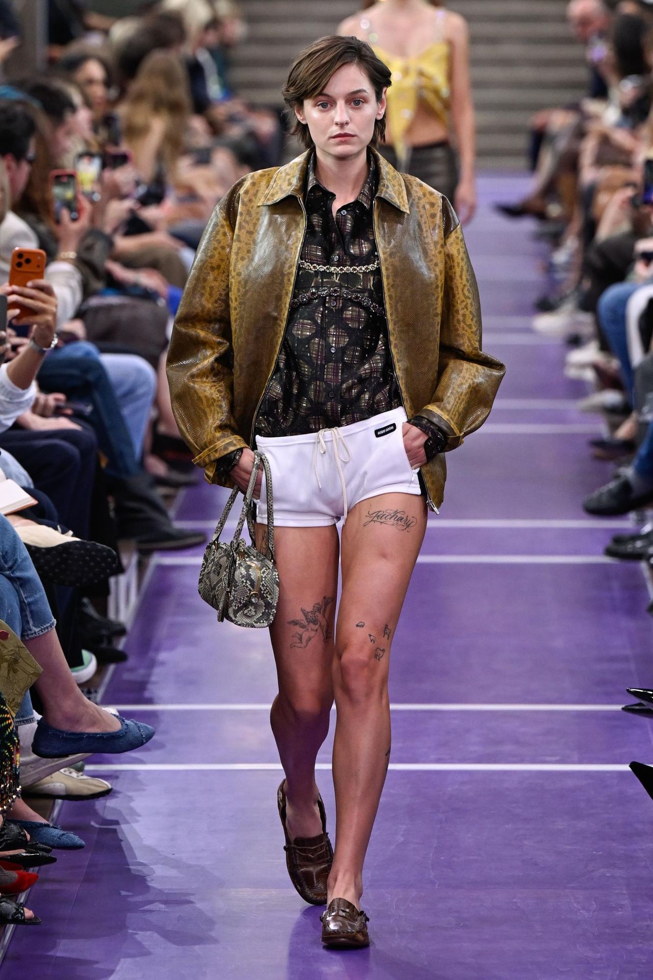 Emma Corrin walks the Miu Miu runway in a textured brown leather jacket layered over a sheer black top, paired with white drawstring shorts, a patterned handbag and brown loafers. | Source: Getty Images