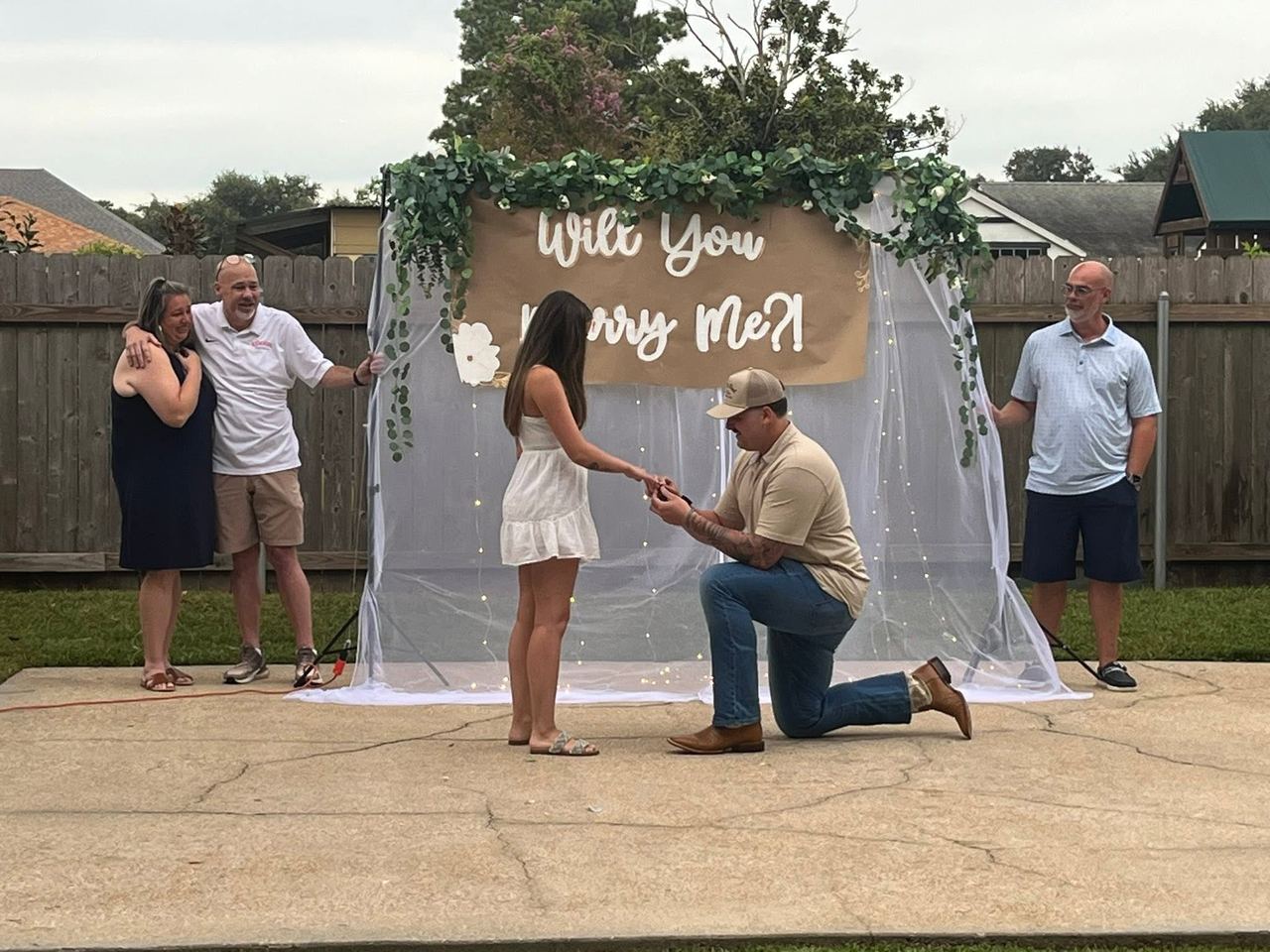 Shane David Hirstius drops to one knee to propose to his longtime love Leah Victoria Fonte during a backyard proposal setup. Wearing a beige collared shirt, jeans, boots, and a cap, Shane slips an engagement ring onto Leah’s finger as she stands before him in a white sundress in front of an arch adorned with greenery, fairy lights, and a banner reading "Will You Marry Me?!", while family members look on emotionally from either side. | Source: Facebook/StevenMarjorie Hirstius