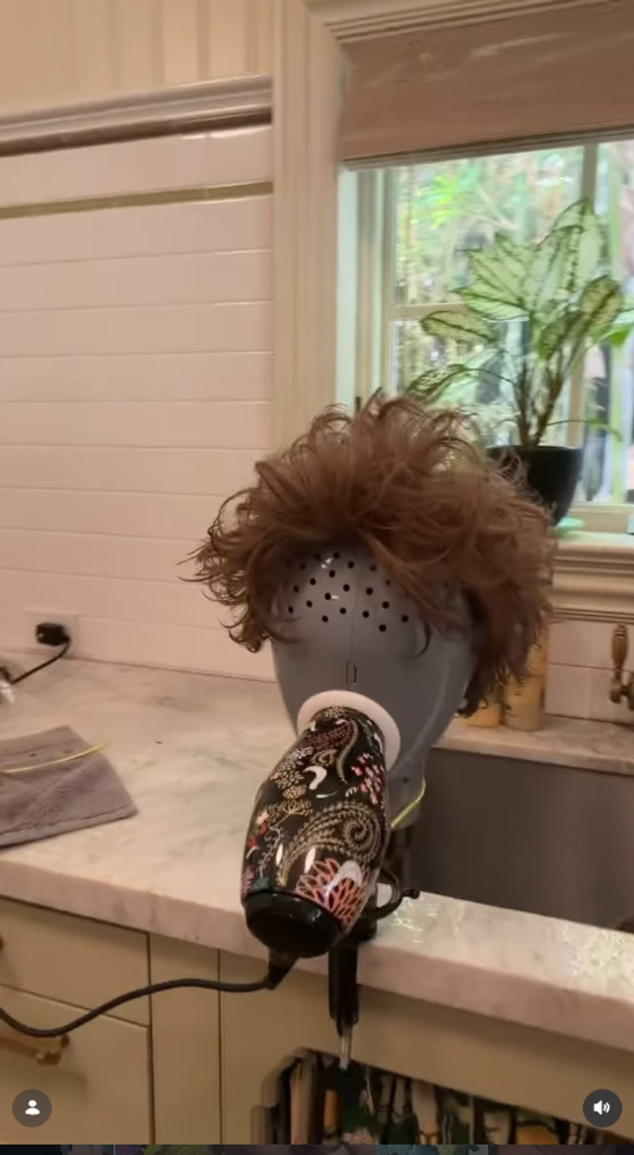 One of McCarthy’s wigs sits on a stand as she uses a hair dryer to dry the freshly washed piece. | Source: Instagram/melissamccarthy