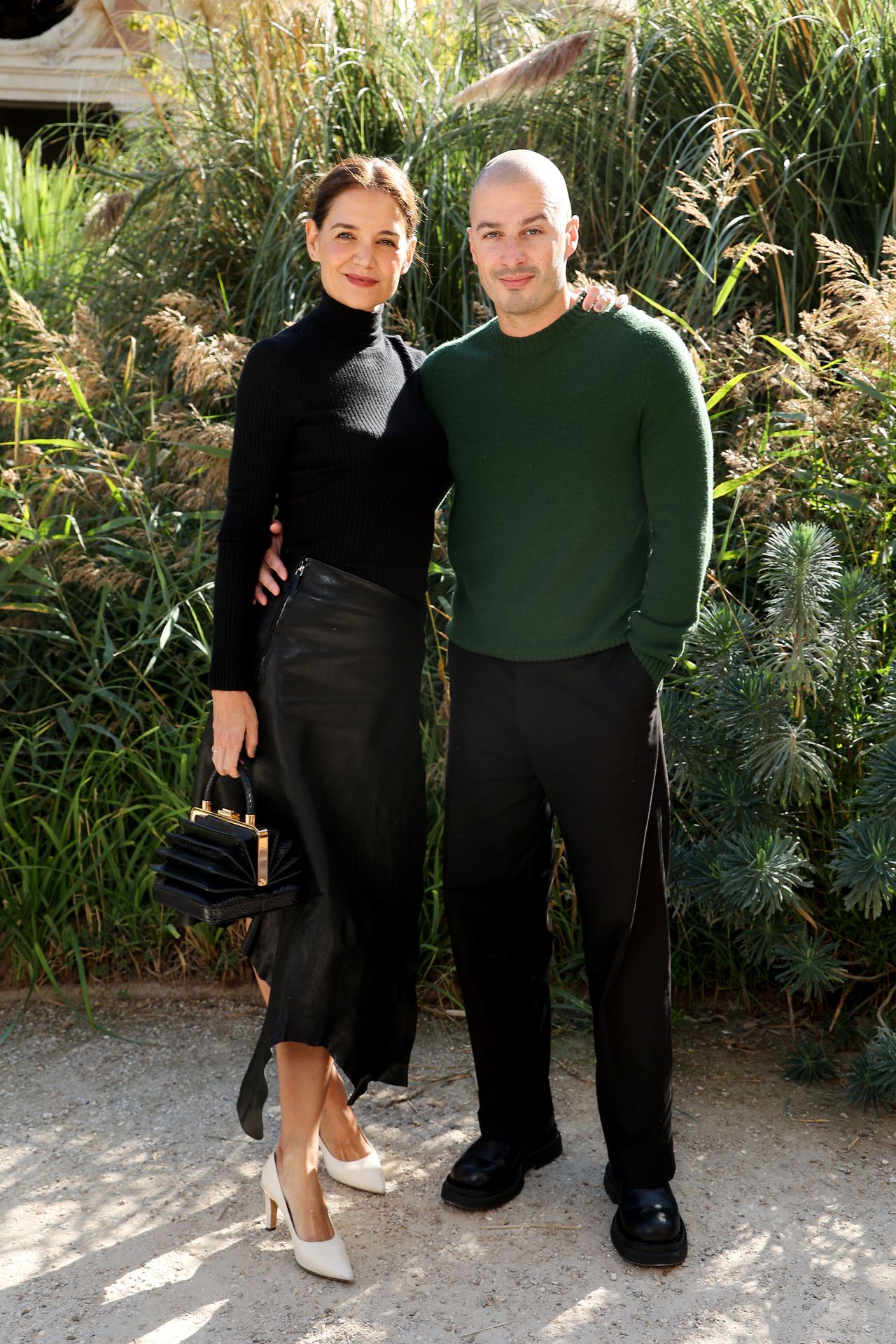 White pointed pumps break up Katie Holmes' otherwise black outfit, while Jason Bard Yarmosky's forest-green sweater adds the strongest color beside her. The actress and artist arrived together for Gabriela Hearst's Spring/Summer 2027 show in Paris. | Source: Getty Images
